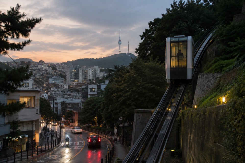 Glass-walled inclined elevator rising along a hillside above a Seoul street