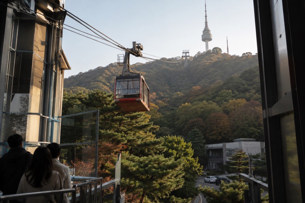 A cable car cabin ascending above a forested ridge toward a tower on the summit