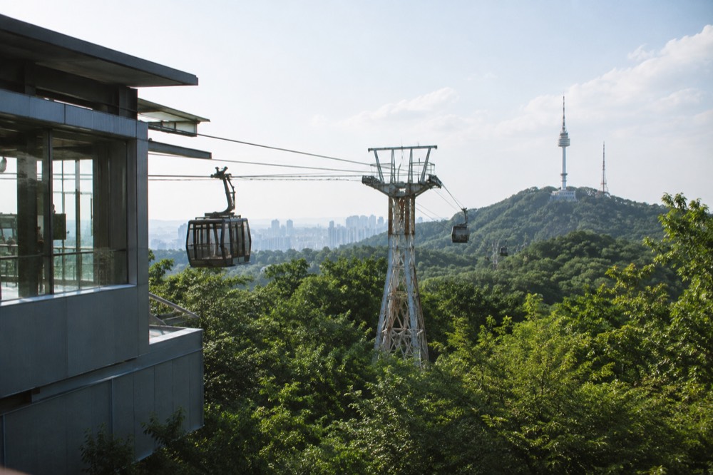 Cable car cabins suspended on the ropeway above dense green forest with Seoul buildings in the distance
