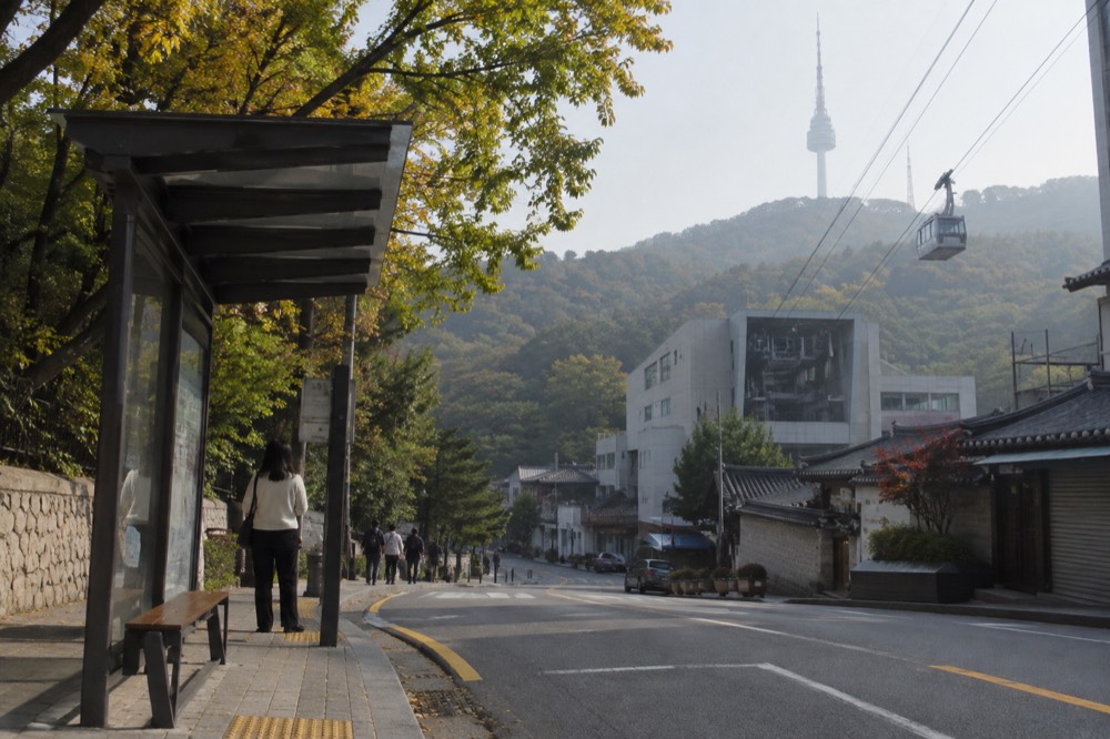A bus stop sign beside a city street with shops and a hillside rising behind the buildings