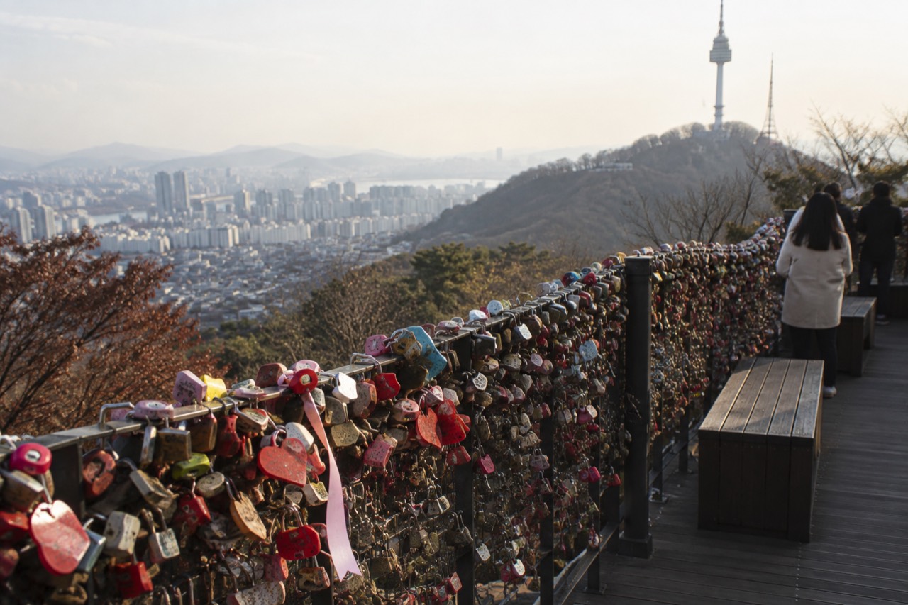 Rows of padlocks attached to a wire fence on a hillside terrace above a city skyline