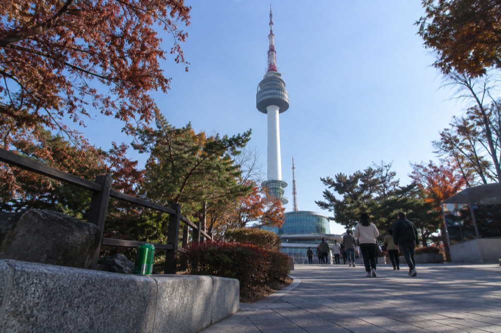Visitors walking on a hilltop plaza toward a tall tower, railings and trees along the path