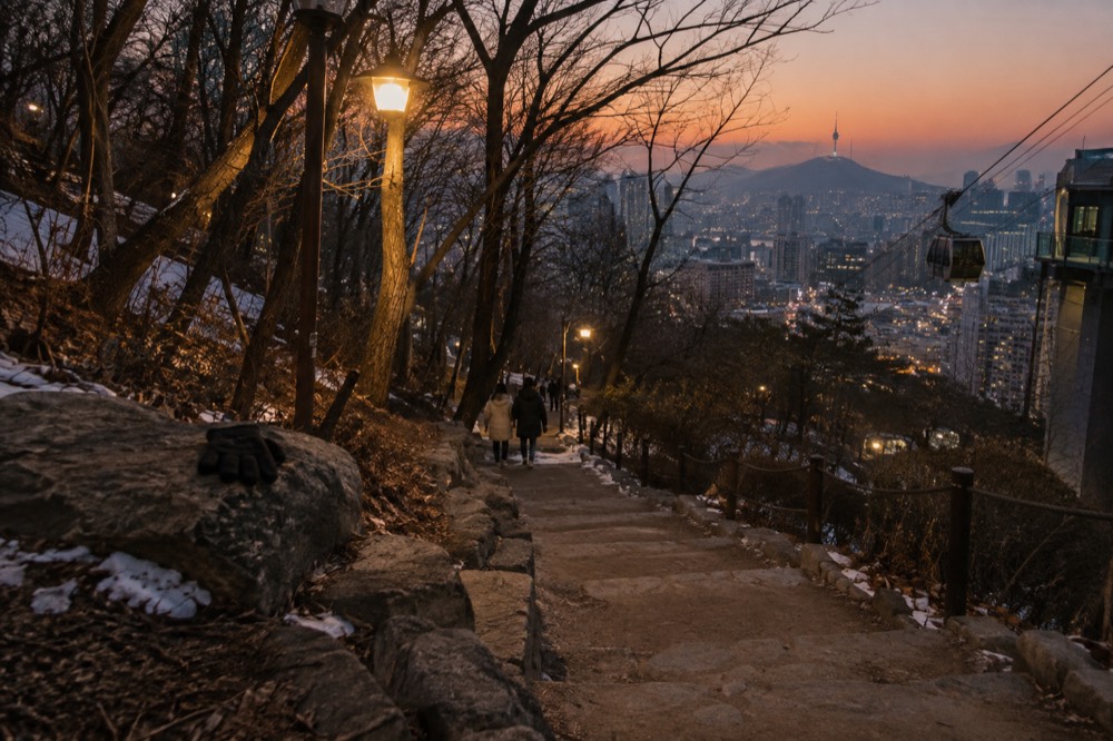 A lit walking trail on Namsan at dusk with the city glowing below